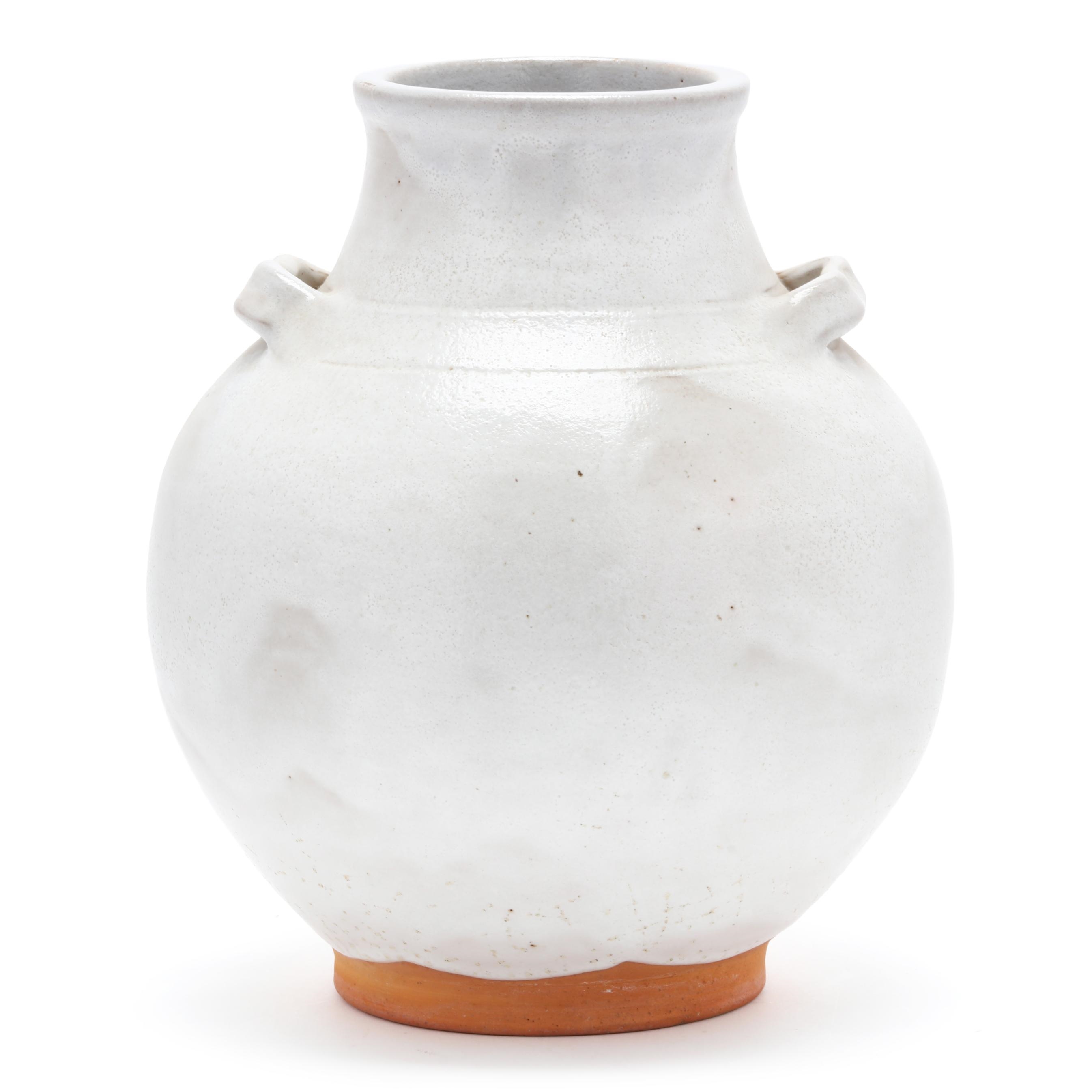 A white ceramic vase with a rounded body and small handles, resting on an exposed terracotta base.