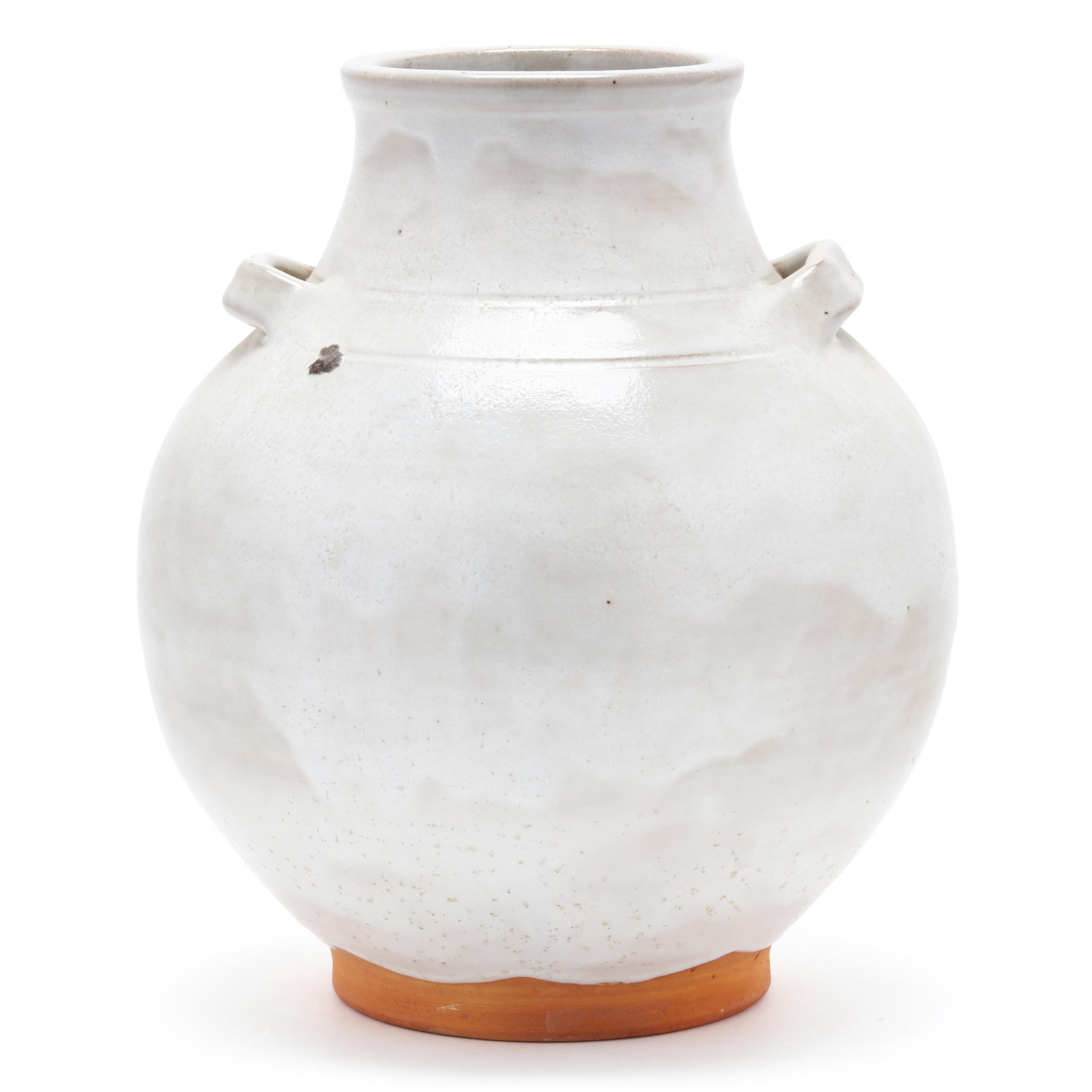A white ceramic vase with a rounded body and small handles, resting on an exposed terracotta base.