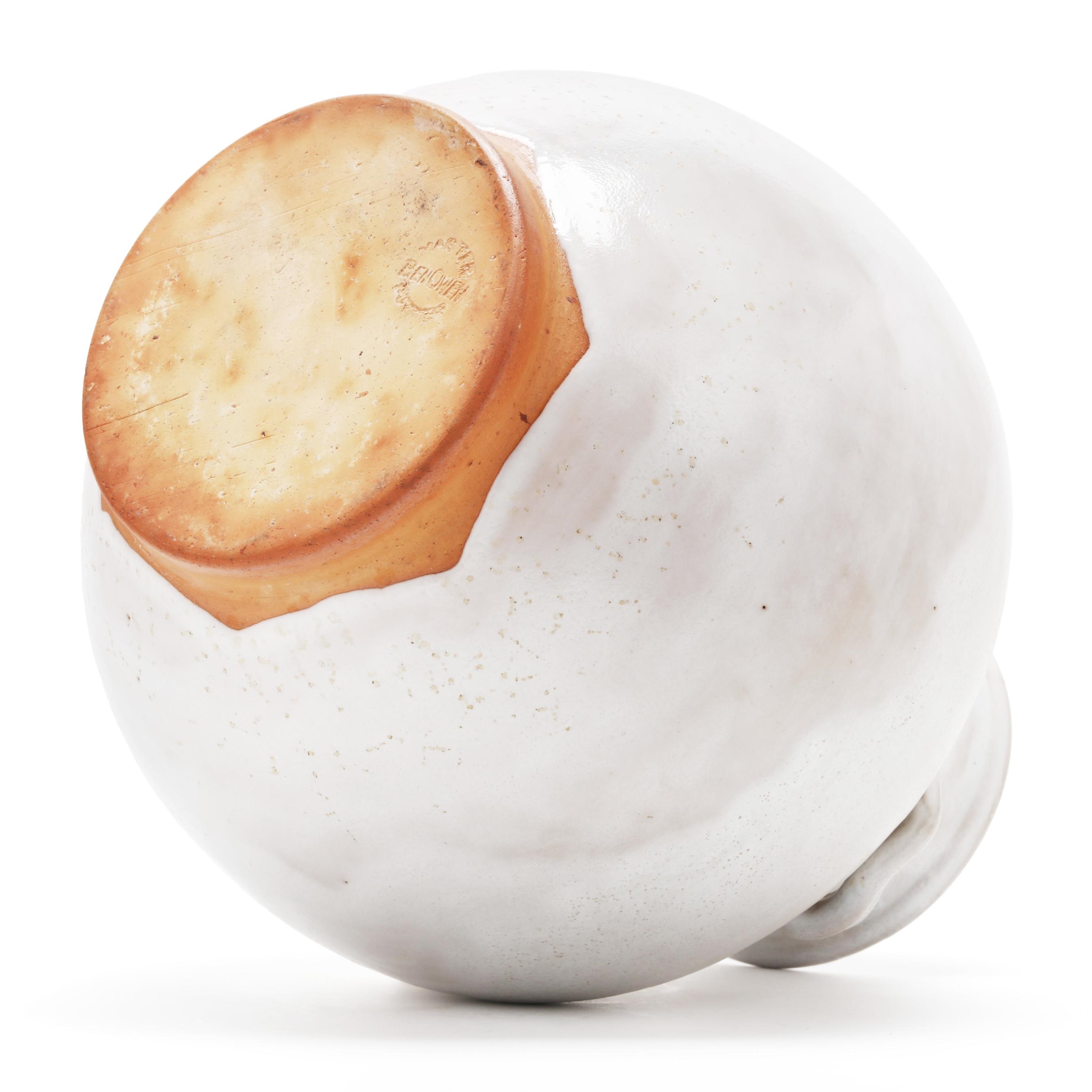 A white ceramic vase with a rounded body and small handles, resting on an exposed terracotta base.