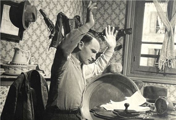 A black-and-white photograph of a man in a domestic interior, arms raised as if caught in a moment of intense emotion or action.