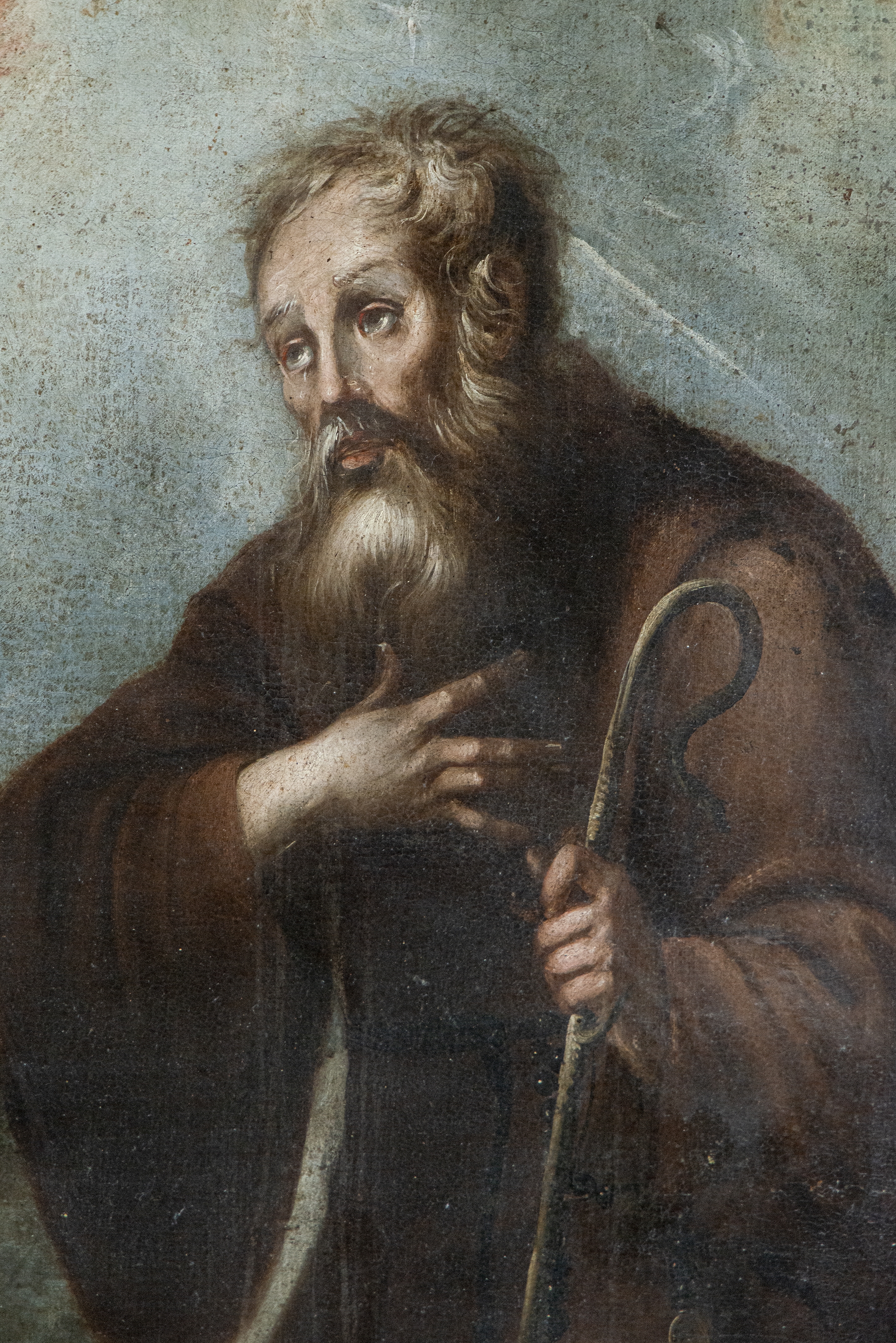 A solemn religious portrait of a bearded figure in dark robes holding a staff, set against a dramatic cloudy sky.