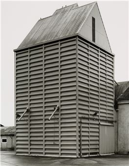 A black-and-white architectural photograph of a tall, slatted industrial building with a peaked roof.