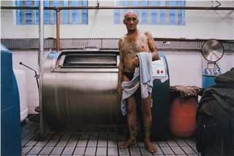 A tattooed figure stands shirtless in a laundry room, holding a towel near an industrial washing machine.