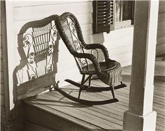 A vintage wicker rocking chair casts an intricate shadow on a wooden porch wall.