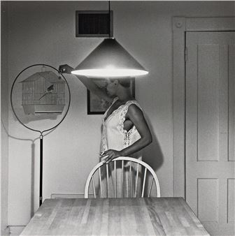 A black-and-white photograph of a person reaching for a hanging lamp while holding a birdcage.