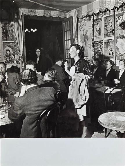 A black-and-white photograph of a lively café scene where a woman stands addressing seated patrons.
