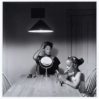 A black-and-white photograph captures a woman and a young girl applying makeup at a dining table under a single pendant light.