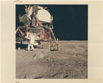 A lone figure in a bulky white suit stands beside a large, complex mechanical structure on a barren, cratered surface under a black sky.