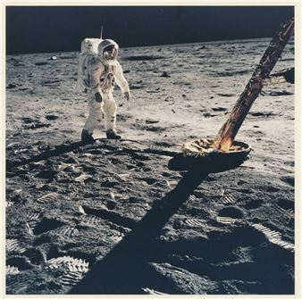 A figure in a bulky white suit stands on a barren, cratered surface next to a rusted metal structure.