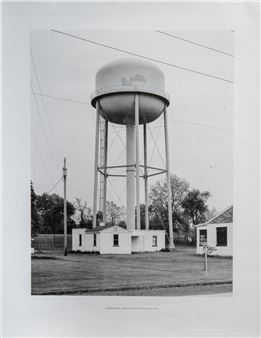 A black-and-white photograph of a water tower standing over a quiet rural landscape.