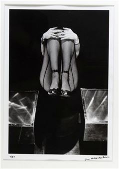 A black-and-white portrait of a seated figure with crossed legs and hands covering the face.