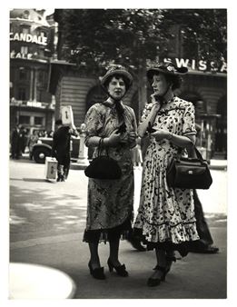 Two women in vintage dresses converse on a city street.