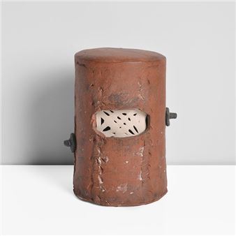 A rusted metal cylinder with a perforated face and bolts, resembling a stylized helmet or industrial object.