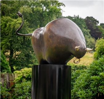 A bronze bull sculpture with a coiled tail and gold-tipped horns stands on a black pedestal against a lush green background.