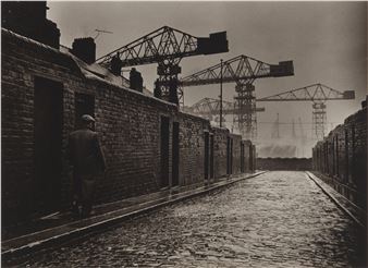 A solitary figure walks along a brick-lined canal path with towering cranes looming in the background.