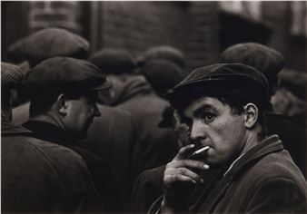 A black-and-white portrait of a man smoking in a crowd, capturing a moment of quiet intensity.