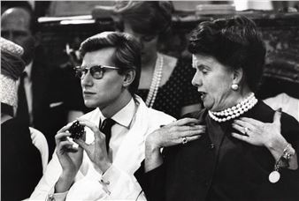 A black-and-white portrait captures a man in sunglasses holding a small object while a woman gestures toward her neck.