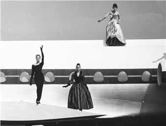 A black-and-white fashion photograph featuring three figures in dramatic poses on a stylized runway.