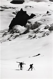 A high-contrast black and white photograph of two skiers ascending a vast, snow-covered mountain.