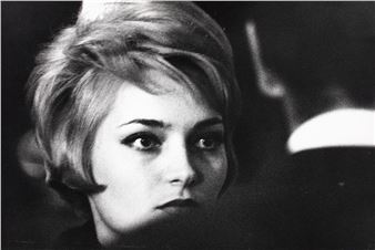 A black-and-white portrait of a person with short hair and dramatic makeup, gazing forward against a dark background.