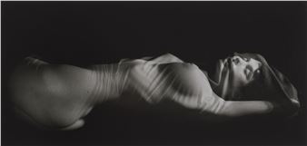 A dramatic black and white photograph of a nude figure lying on their back with light casting sharp shadows across the body.