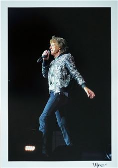 A performer on stage captured mid-song, wearing a patterned jacket and jeans.