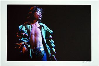 A dramatic performance photograph capturing a shirtless figure in a ruffled green jacket mid-song.