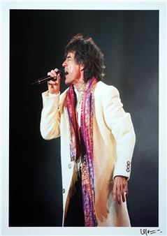 A performer captured mid-song on a dark stage, wearing a white coat and colorful scarf.
