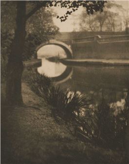 A sepia-toned photograph of a stone bridge arching over a calm canal, framed by a tree trunk and reflecting water.