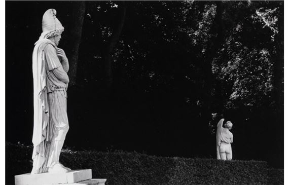 A black-and-white photograph featuring two classical statues in a dark, wooded setting.