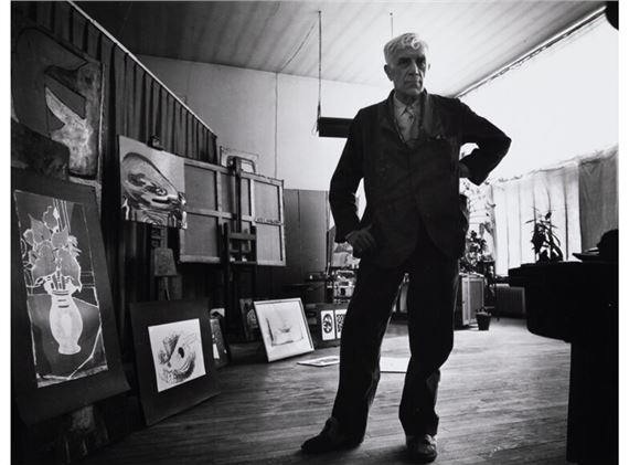 A black-and-white portrait of a man standing confidently in an art studio filled with framed drawings.