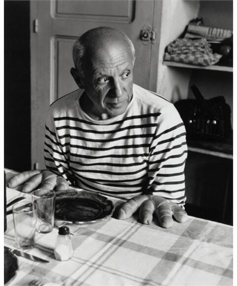 A black-and-white portrait of a man in a striped shirt leaning over a table with bread and plates.