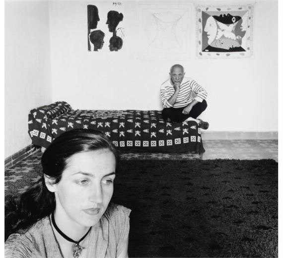 A black-and-white portrait of a woman in the foreground with a man sitting on a bed in the background, framed by patterned textiles and wall art.