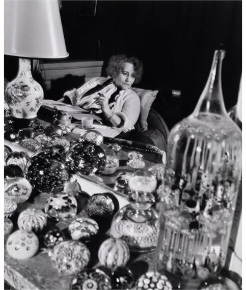 A person sits at a table surrounded by numerous glass ornaments and decorative objects.