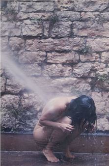 A person crouches under a shower against a rough stone wall.