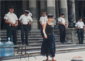 A solitary figure stands barefoot in front of a formal building, surrounded by uniformed personnel.
