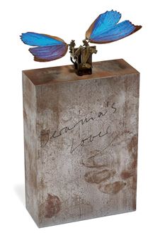 A sculptural piece featuring a metallic block with handwritten text and a small figure adorned with large blue butterfly wings.