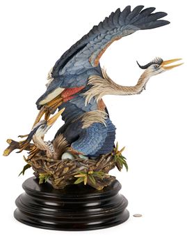 A detailed sculptural piece featuring a large heron with wings spread over a nest containing two smaller birds.