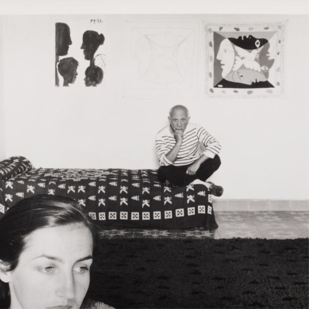 A black-and-white interior scene featuring a woman in the foreground and a man sitting on a patterned sofa in the background.