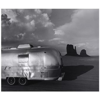 A vintage metallic trailer parked in a vast desert landscape with dramatic rock formations under a cloudy sky.