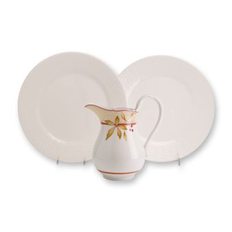 A white ceramic pitcher with autumnal leaf details sits between two textured plates on a plain background.