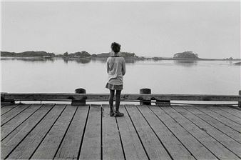 A solitary figure stands on a wooden pier overlooking a calm body of water.