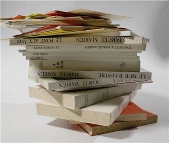 A precarious stack of hardcover books with visible spines, leaning against a plain background.