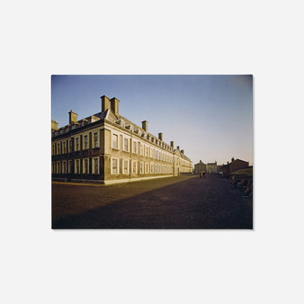 "A long, multi-story building with a row of chimneys stands under a clear blue sky, casting a long shadow across the ground."