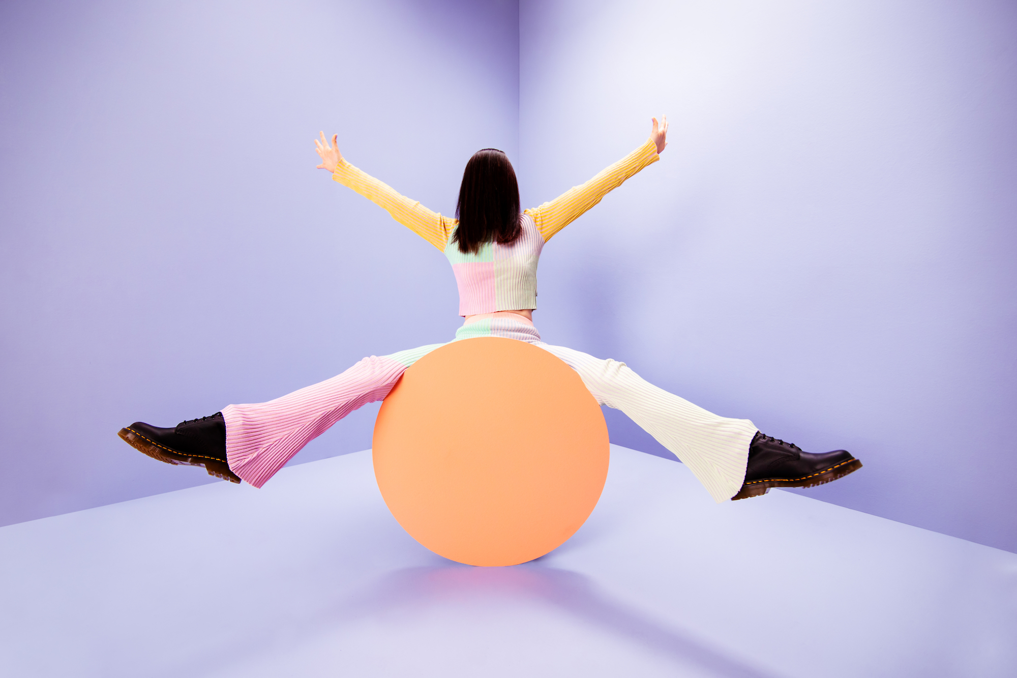 A person in a colorful outfit balances on an orange sphere with arms raised against a purple studio background.