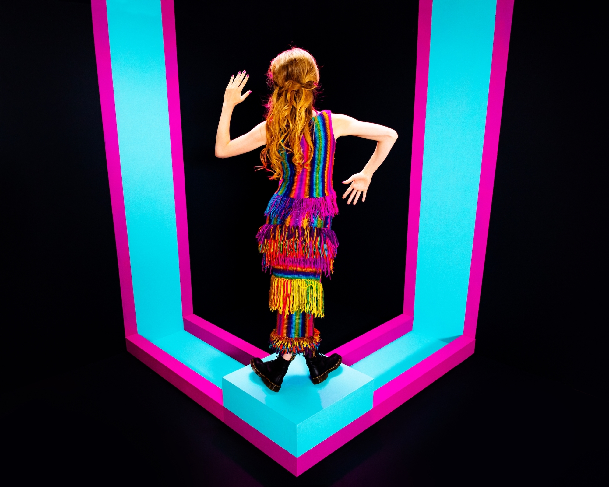 A colorful figure stands inside a neon-lit geometric frame, waving with one hand.
