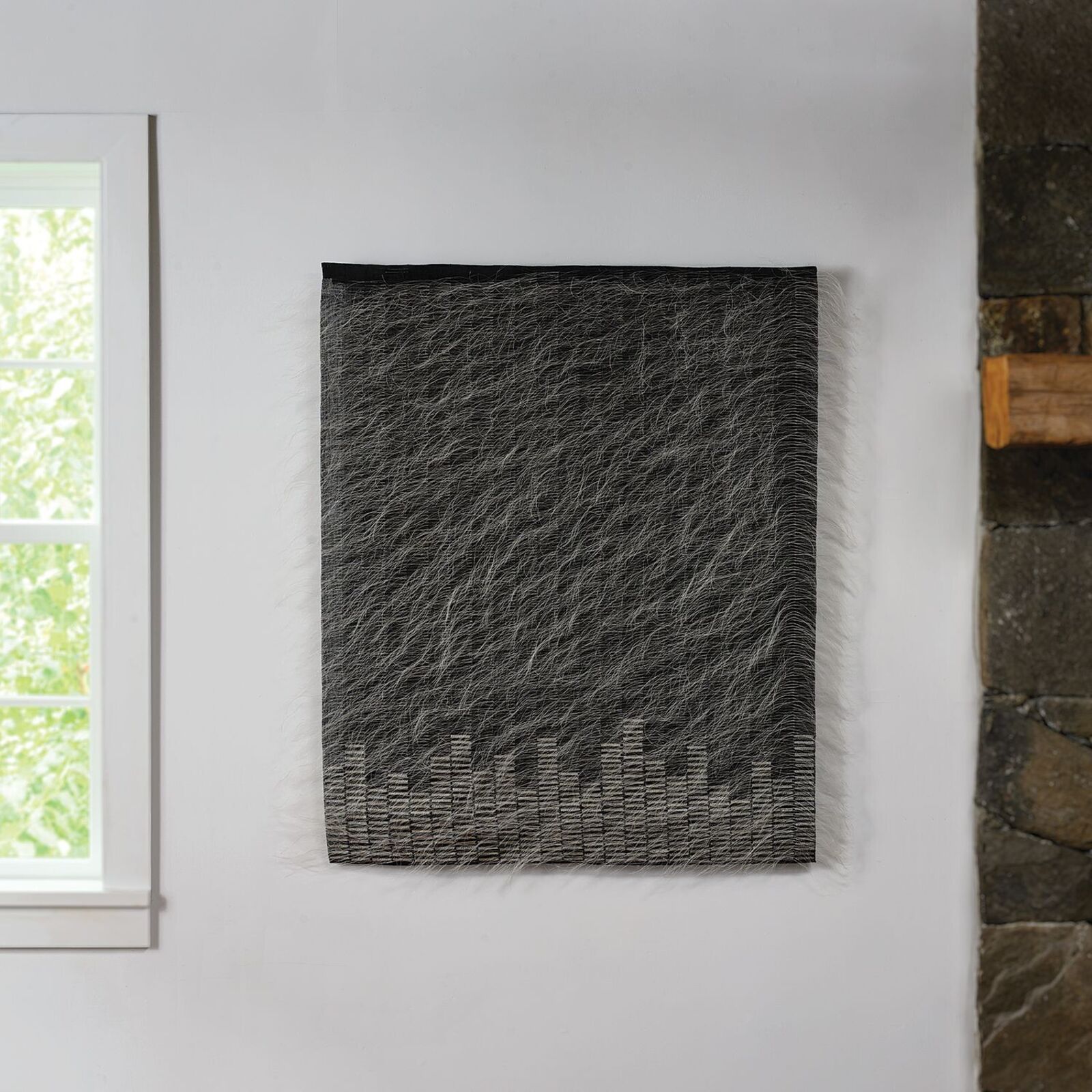 A textured textile artwork featuring a city skyline silhouette at the bottom, displayed on a white wall.