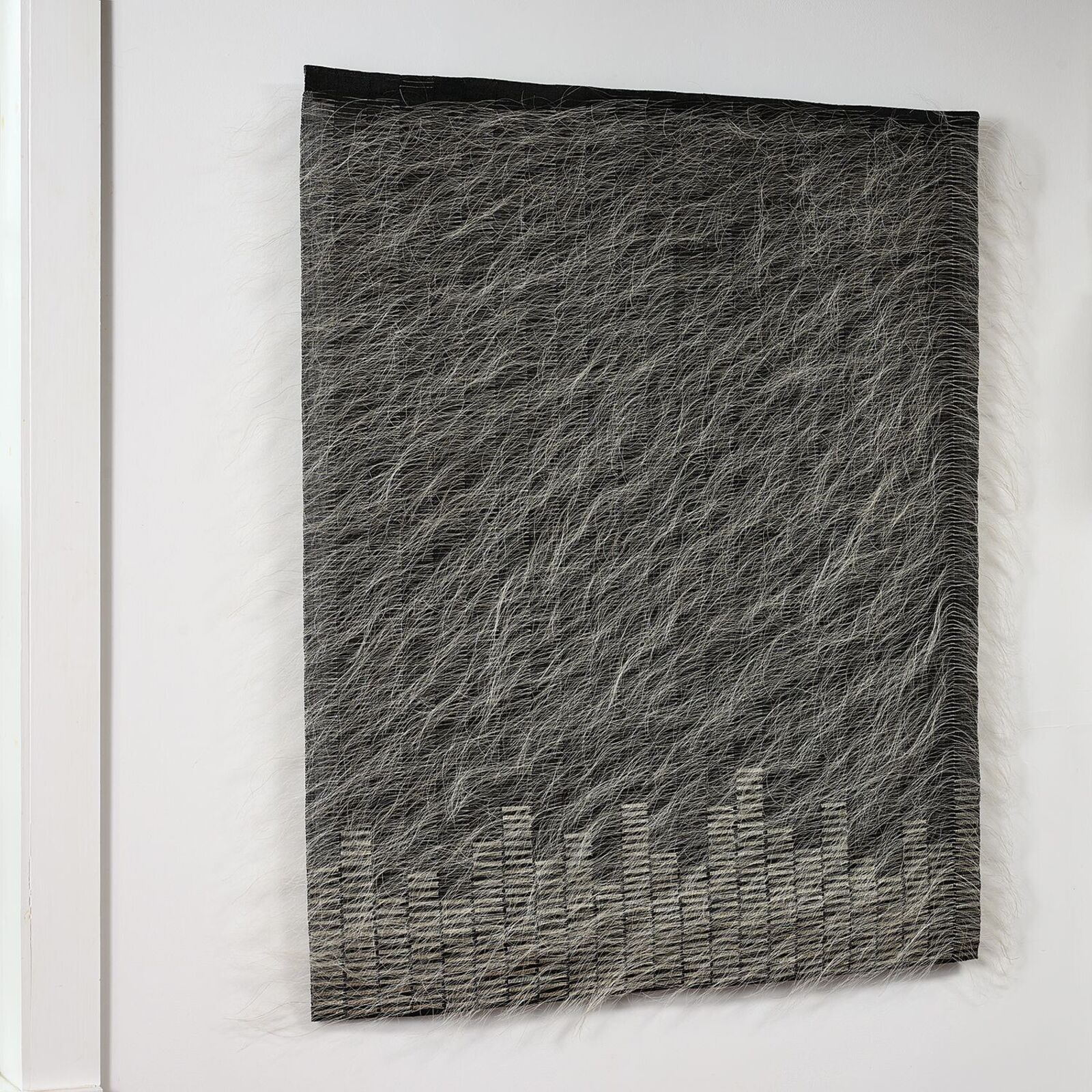 A textured textile artwork featuring a city skyline silhouette at the bottom, displayed on a white wall.