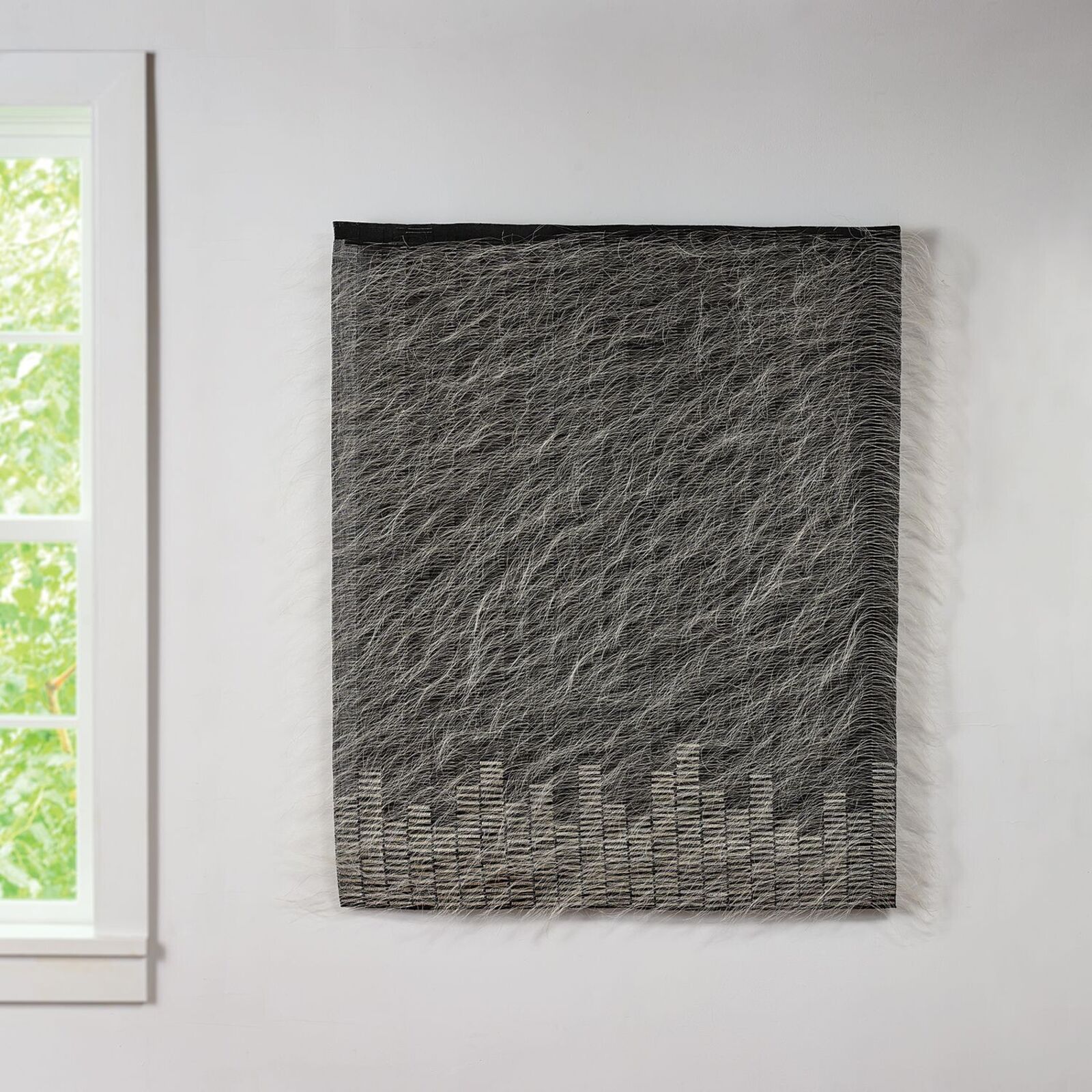 A textured textile artwork featuring a city skyline silhouette at the bottom, displayed on a white wall.