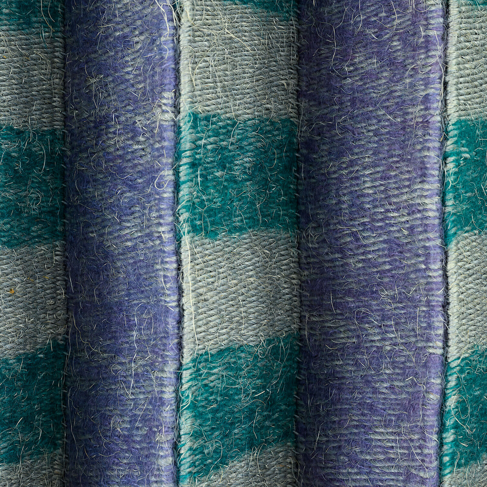A geometric textile wall hanging featuring a pleated, accordion-fold design in teal, purple, and grey tones.